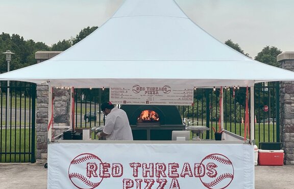 The picture shows a white folding gazebo that serves as a street food stand. The front sidewall on half height is printed with the red company logo. Under the tent there is a pizza oven and the pizza maker.