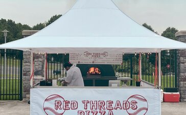The picture shows a white folding gazebo that serves as a street food stand. The front sidewall on half height is printed with the red company logo. Under the tent there is a pizza oven and the pizza maker.