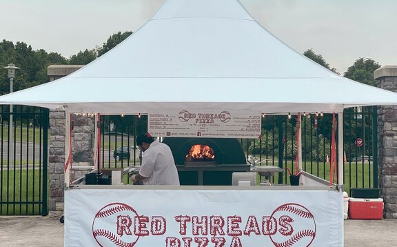 L'immagine mostra un gazebo pieghevole bianco che serve come stand di cibo di strada. La parete laterale anteriore è a metà altezza ed è stampata con il logo rosso dell'azienda. Sotto il gazebo c'è un forno per la pizza e il pizzaiolo..