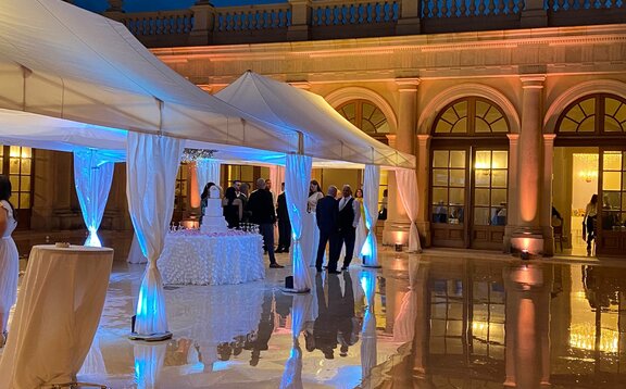 It is evening. On the terrace of a large building are two large, white wedding tents. Below them are several guests and the wedding cake.