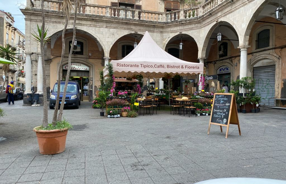On a square in front of a restaurant there is a large 5x5 m folding gazebo with a scalloped valance. Underneath there are tables, chairs and flowers.