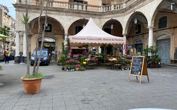 On a square in front of a restaurant there is a large 5x5 m folding gazebo with a scalloped valance. Underneath there are tables, chairs and flowers.