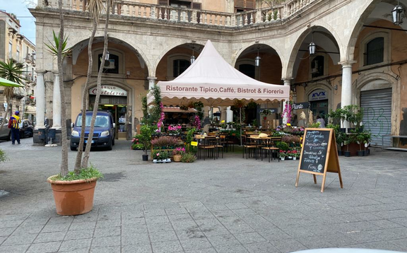 Su una piazza di fronte a un ristorante c'è un grande gazebo pieghevole di 5x5 m con un frontalino ondulato. Sotto ci sono tavoli, sedie e fiori.