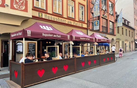 Next to a street in front of a restaurant there are three folding gazebos with red roofs. Under the tent there are tables and benches.