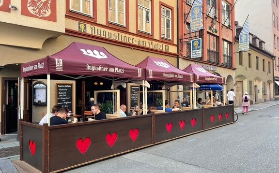 Next to a street in front of a restaurant there are three folding gazebos with red roofs. Under the tent there are tables and benches.