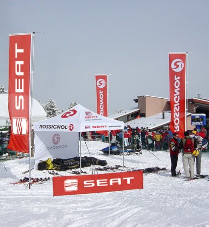 In a ski resort there is a 3x3 m folding gazebo with a white roof and a white sidewall printed in red. There are three large red flags around the gazebo. In the background there are several skiers and buildings.