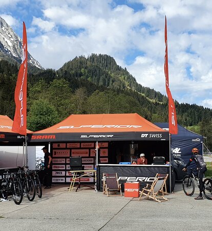 Es stehen zwei Faltpavillons mit orangen Dächern und Flaggen auf einem Platz, rechts daneben ein Wohnwagen. Im Hintergrund befinden sich Wälder und Berge.