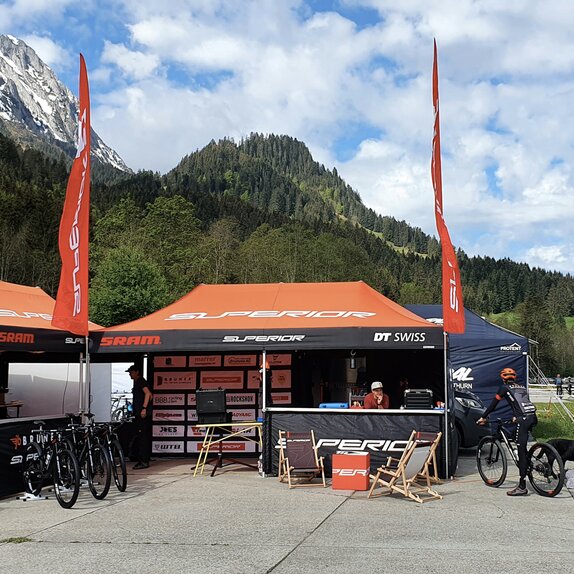 Es stehen zwei Faltpavillons mit orangen Dächern und Flaggen auf einem Platz, rechts daneben ein Wohnwagen. Im Hintergrund befinden sich Wälder und Berge.