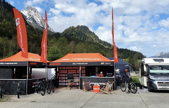 Es stehen zwei Faltpavillons mit orangen Dächern und Flaggen auf einem Platz, rechts daneben ein Wohnwagen. Im Hintergrund befinden sich Wälder und Berge.