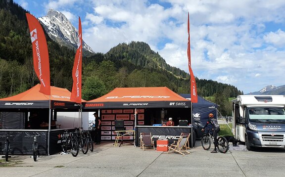 Es stehen zwei Faltpavillons mit orangen Dächern und Flaggen auf einem Platz, rechts daneben ein Wohnwagen. Im Hintergrund befinden sich Wälder und Berge.