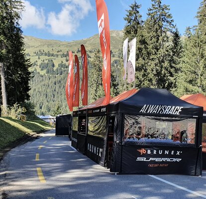On a road there are several black folding gazebos with orange flags and roofs of a racing team. The road is surrounded by woods.