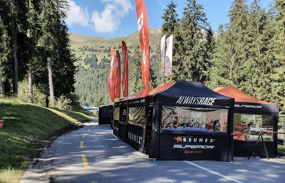 On a road there are several black folding gazebos with orange flags and roofs of a racing team. The road is surrounded by woods.