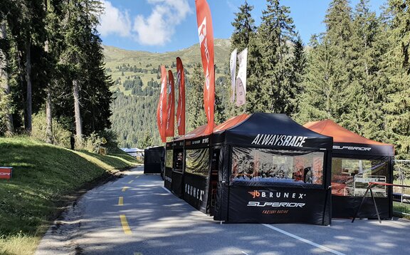 Su una strada ci sono diversi gazebo pieghevoli neri con bandiere e tetti arancioni di una squadra da corsa. La strada è circondata da boschi.