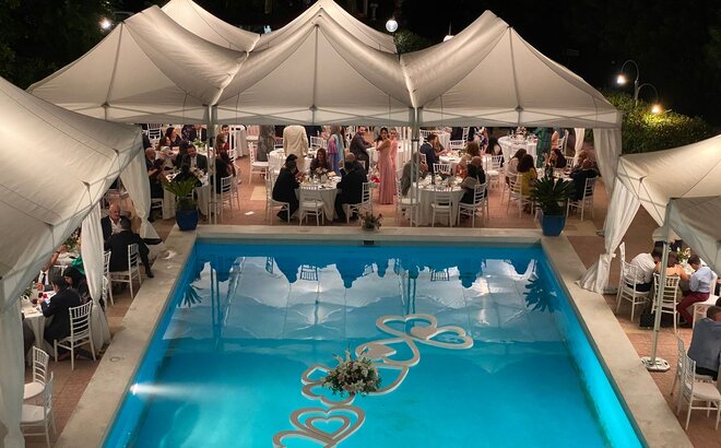 Elegant wedding tents in white stand by the pool. Underneath there are many people sitting at tables. The elegant corner curtains add a special flair.