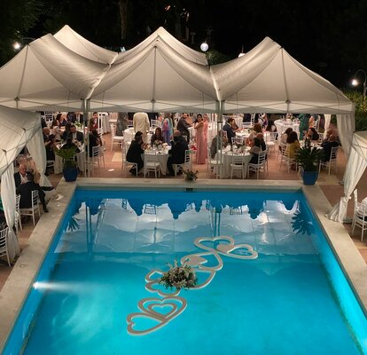 Elegante Hochzeitszelte in weiß stehen am Pool. Darunter sitzen zahlreiche Personen an Tischen. Die eleganten Eckvorhänge verleihen ein besonderes Flair.