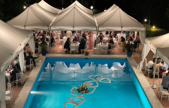 Elegant wedding tents in white stand by the pool. Underneath there are many people sitting at tables. The elegant corner curtains add a special flair.