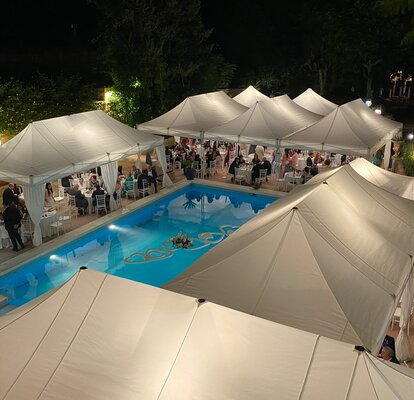 Elegante Hochzeitszelte in weiß stehen am Pool. Darunter sitzen zahlreiche Personen an Tischen. Die eleganten Eckvorhänge verleihen ein besonderes Flair.