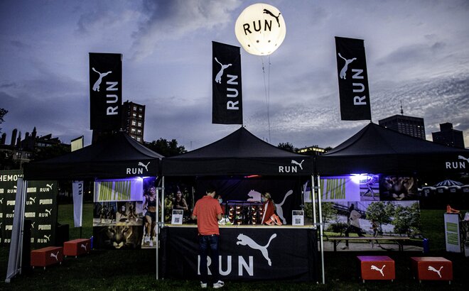 Three 3x3 m folding gazebos are set up side by side on a lawn at a sports event. The folding gazebos are black and personalized with the Puma logo. All three also have a personalized flag on the roof.