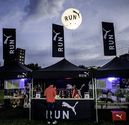 Three 3x3 m folding gazebos are set up side by side on a lawn at a sports event. The folding gazebos are black and personalized with the Puma logo. All three also have a personalized flag on the roof.