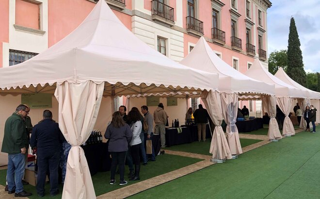 There are four white folding gazebos in front of a building. There are many people under and next to the tents, a wine tasting is taking place.