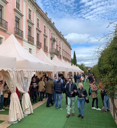 Hay diez carpas plegables blancas delante de un edificio. Hay mucha gente debajo y al lado de las carpas, se está realizando una cata de vinos.