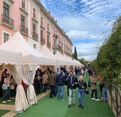Hay diez carpas plegables blancas delante de un edificio. Hay mucha gente debajo y al lado de las carpas, se está realizando una cata de vinos.