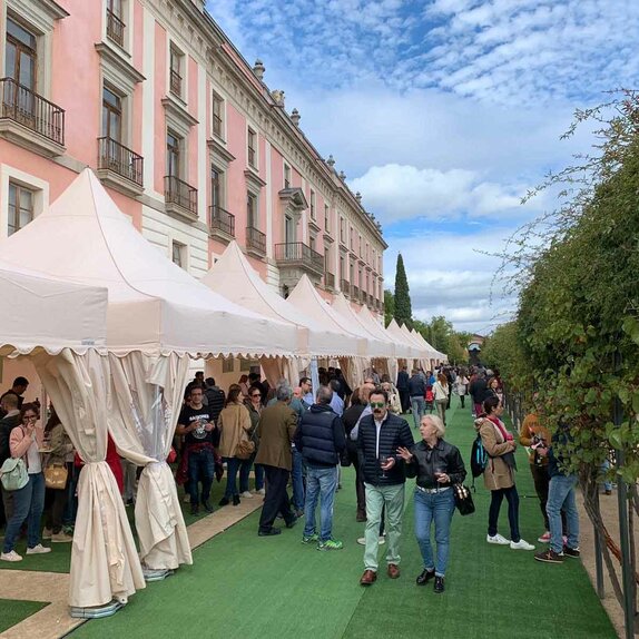 Hay diez carpas plegables blancas delante de un edificio. Hay mucha gente debajo y al lado de las carpas, se está realizando una cata de vinos.