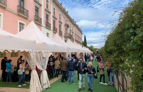 Hay diez carpas plegables blancas delante de un edificio. Hay mucha gente debajo y al lado de las carpas, se está realizando una cata de vinos.
