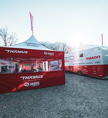 Sulla sinistra dell'immagine si vede un gazebo pieghevole di 5x5 m con tetto bianco e pareti laterali rosse. Le pareti laterali sono dotate di un'ampia finestra panoramica in PVC e il gazebo dispone anche di una bandiera sul tetto. A sua destra si trova un grande camper.