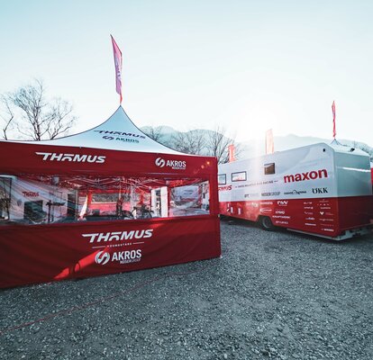 Sulla sinistra dell'immagine si vede un gazebo pieghevole di 5x5 m con tetto bianco e pareti laterali rosse. Le pareti laterali sono dotate di un'ampia finestra panoramica in PVC e il gazebo dispone anche di una bandiera sul tetto. A sua destra si trova un grande camper.