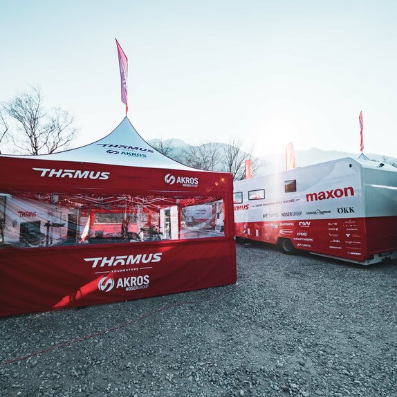 Sulla sinistra dell'immagine si vede un gazebo pieghevole di 5x5 m con tetto bianco e pareti laterali rosse. Le pareti laterali sono dotate di un'ampia finestra panoramica in PVC e il gazebo dispone anche di una bandiera sul tetto. A sua destra si trova un grande camper.