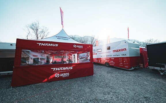 Sulla sinistra dell'immagine si vede un gazebo pieghevole di 5x5 m con tetto bianco e pareti laterali rosse. Le pareti laterali sono dotate di un'ampia finestra panoramica in PVC e il gazebo dispone anche di una bandiera sul tetto. A sua destra si trova un grande camper.