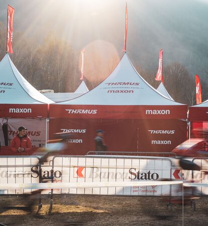 The picture shows three folding gazebos with red roof flags of the Thömus Maxon mountain bike team. The tents have white roofs and red sidewalls. In the background you can see three more roof peaks with the flags.