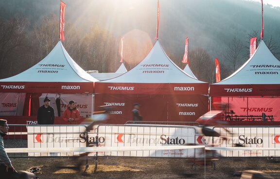 The picture shows three folding gazebos with red roof flags of the Thömus Maxon mountain bike team. The tents have white roofs and red sidewalls. In the background you can see three more roof peaks with the flags.