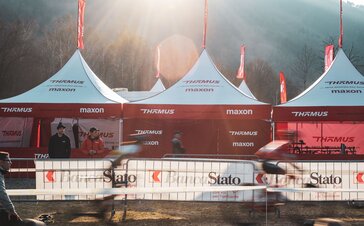 The picture shows three folding gazebos with red roof flags of the Thömus Maxon mountain bike team. The tents have white roofs and red sidewalls. In the background you can see three more roof peaks with the flags.