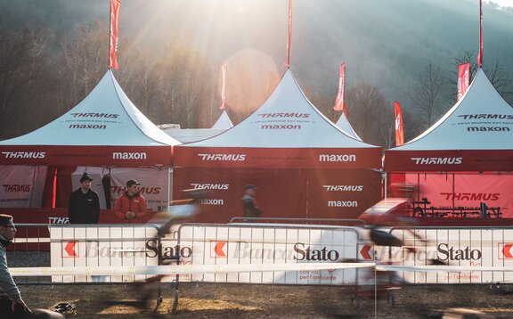 L'immagine mostra tre gazebo pieghevoli con bandiere rosse sul tetto della squadra di mountain bike Thömus Maxon. Hanno il tetto bianco e le pareti laterali rosse. Sullo sfondo si vedono altri tre tetti con le bandiere.