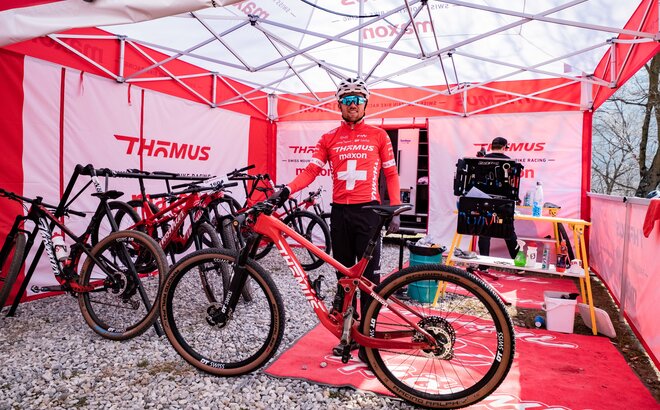 The photo shows a mountain bike racer with his bike. He is standing under a red and white 5x5 m folding gazebo. There are also many other bikes and a second person under the tent.