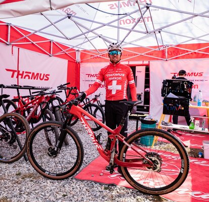 La foto mostra un corridore di mountain bike con la sua bicicletta. È sotto un gazebo pieghevole rosso e bianco di 5x5 metri. Ci sono anche molte altre biciclette e una seconda persona sotto il gazebo.