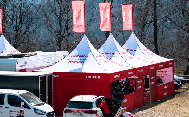 The picture shows three big folding gazebos with red roof flags of the Thömus Maxon mountain bike team. The tents have white roofs and red sidewalls with windows and doors. There are also vehicles and people in the picture.