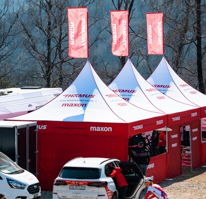 The picture shows three big folding gazebos with red roof flags of the Thömus Maxon mountain bike team. The tents have white roofs and red sidewalls with windows and doors. There are also vehicles and people in the picture.