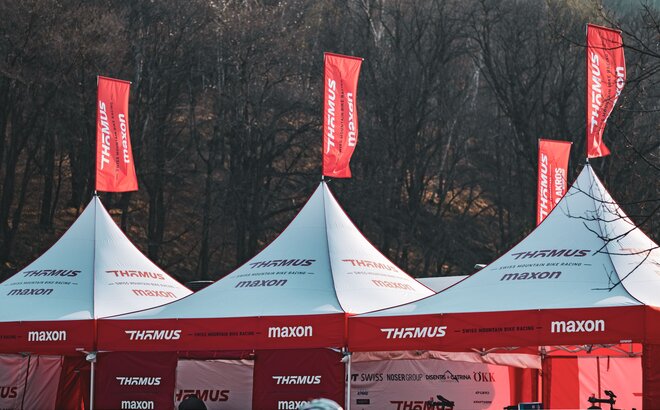 The picture shows three folding gazebos with red roof flags of the Thömus Maxon mountain bike team. The tents have white roofs and red sidewalls. A large hill with trees can be seen in the background.