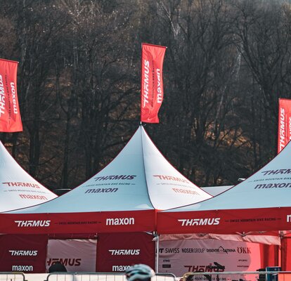 L'immagine mostra due gazebo pieghevoli con bandiere rosse sul tetto della squadra di mountain bike Thömus Maxon. Hanno il tetto bianco e le pareti laterali rosse. Sullo sfondo si vede una grande collina con alberi.