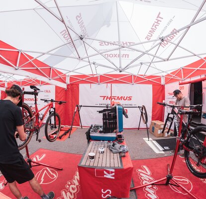 Under a folding gazebo, two men who are repairing mountain bikes can be seen.