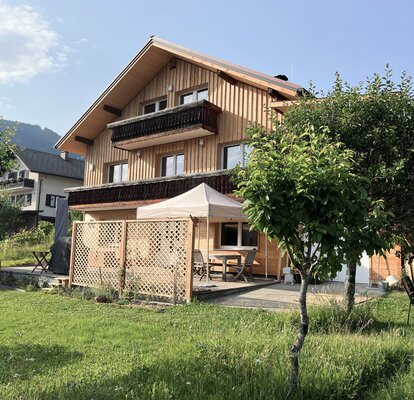 Auf einer Wiese steht ein Haus mit einer Terrasse. Auf der Terrasse befindet sich ein beiges Faltzelt, das mit Gewichten beschwert ist.