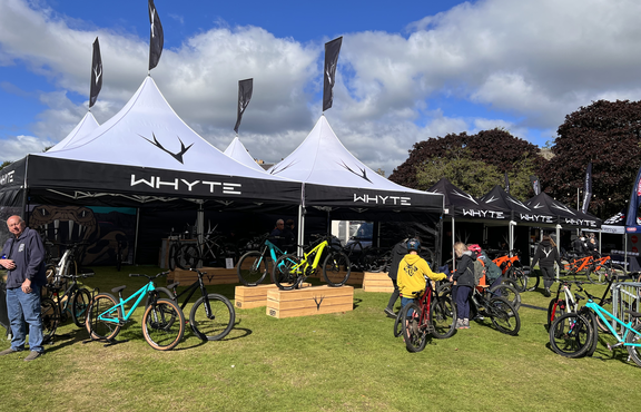 Four 5x5 m folding gazebos with white pagoda roofs are standing on a meadow. They all have a personalised flag on the top of the roof. Below and next to them are bicycles and to the right are more personalised black folding gazebos.