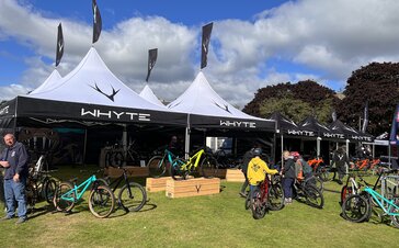 Four 5x5 m folding gazebos with white pagoda roofs are standing on a meadow. They all have a personalised flag on the top of the roof. Below and next to them are bicycles and to the right are more personalised black folding gazebos.