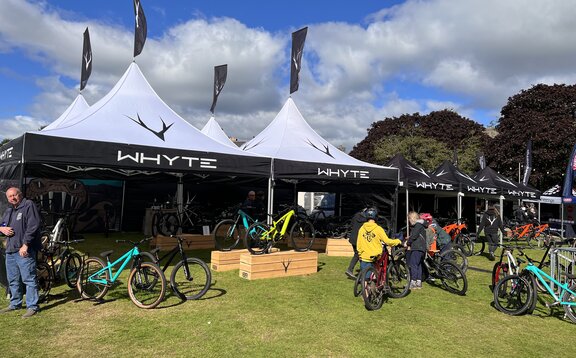 En un prado hay cuatro carpas plegables de 5x5 m con tejados de pagoda blanca. Todos tienen una bandera personalizada encima de los tejados. Debajo y junto a ellos hay bicicletas y, a su derecha, más carpas plegables negras y personalizadas.