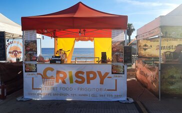 The picture shows a 3x3 m folding gazebo used as a street food vending tent. It has an orange roof, a counter, a sidewall with a door, and is personalized with a promotional print.