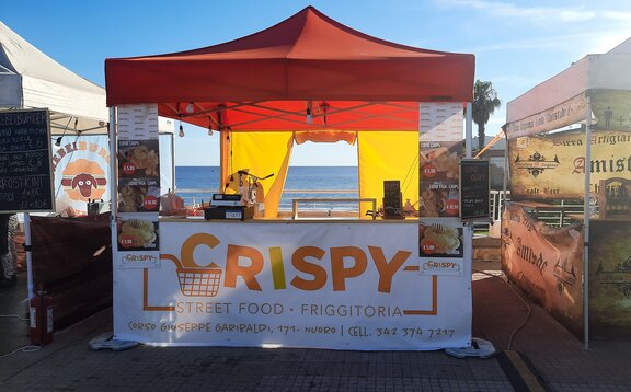 Sur l'image, on peut voir une tente pliante de 3x3 m utilisée comme stand de vente de street food. Elle a un toit orange, un comptoir, une paroi latérale avec une porte et est personnalisée avec une impression publicitaire.