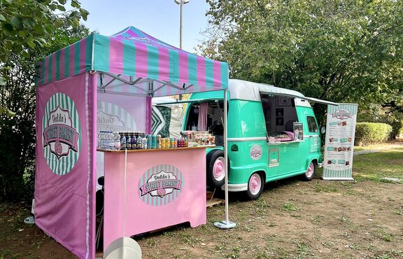 In the picture, there is a 2x2 m large, personalized folding gazebo on a meadow, next to a street food truck. The folding gazebo has three sidewalls printed in pink and blue, just like the roof. It is secured to the ground with weights.